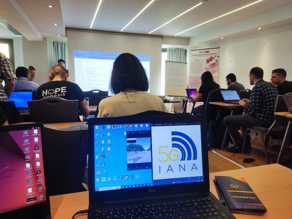 a laptop is on a table, and it shows the 5G-IANA project logo. There are more people in the room sitting on tables and looking at the projector. 